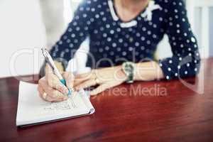 Crossing off tasks from her to do list. Shot of a woman writing in a book at a table.