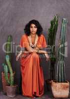 Ola amigos. Studio portrait of a beautiful young ethnic woman sitting amongst cacti against a gray background.