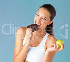 My diet makes room for the occasional indulgence. Studio shot of a fit young woman holding an apple while taking a bite of chocolate against a blue background.