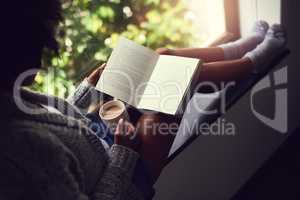 Escaping in the narrative. Shot of an unidentifiable young woman reading a book while enjoying a cup of coffee at home.