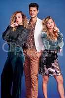 Were off to a 80s themed party. Shot of three young people posing together in 80s clothing against a blue background.