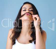 Its all part of her charm. Studio shot of an attractive young woman playfully making a mustache with her hair against a blue background.