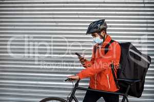 Checking his app for the next delivery location. Shot of a masked man using his cellphone while out on his bicycle for a delivery.