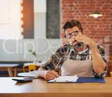 This admin will be the end of me. Shot of an exhausted young business owner doing admin at a table in his coffee shop.