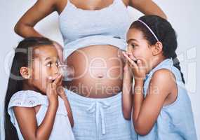 Theres movement in there. Shot of two cheerful little girls standing next their mother while each putting a glass on her pregnant belly to listen for movement at home.