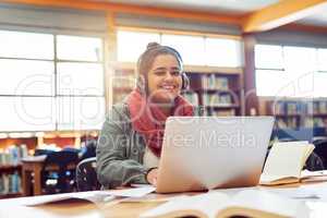 Music makes her perform. Portrait of a cheerful young female student working on a laptop while listening to music with her headphones inside of a library.