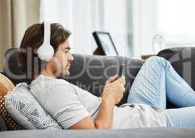 Sending a risky text has never been this nerve wracking. Shot of a young man relaxing on his sofa in the lounge while using his phone and headphones.