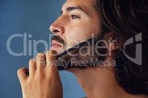 Taming his mane. Studio shot of a handsome young man grooming his facial hair against a blue background.