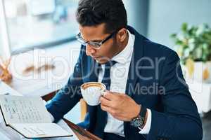 His daily schedule is always full. Shot of a focused young businessman seated at his desk while drinking coffee and reading his journal in the office.