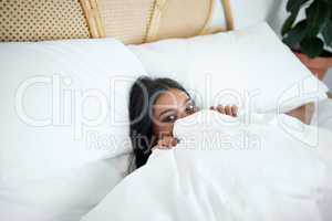 Dont tell anyone that Im here. Shot of a young woman peeking from under her duvet while lying in her bed.