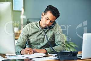 He is constantly busy dealing with clients. Shot of a focused young businessman seated at his desk while taking on the phone and making notes inside the office.
