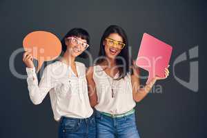 We always think the same. Studio shot of two cheerful young women holding up speech bubbles while standing against a dark background.