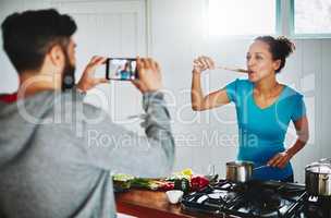 All of the taste, none of the calories. Shot of a man filming his wife on his smartphone while she cooks in their kitchen at home.