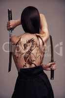 The girl with the dragon tattoo. Conceptual shot of a young female martial artist wielding two blades with a dragon tattoo on her back.