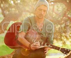 Lyrics inspired by nature. Shot of a handsome young man sitting in a park with a guitar writing song on a notepad.