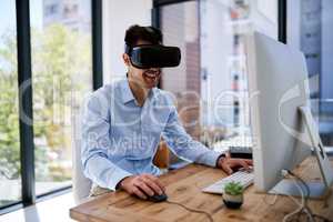 Someday he wont need a screen at all. Shot of a happy young businessman wearing a virtual reality headset while working at his desk in the office.