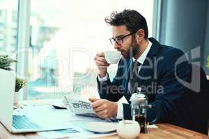 Lets see how the markets are doing. Shot of a young businessman drinking a cup of tea while reading a newspaper in an office.