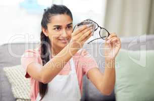 All the dirt is gone and I can see clearly again. Shot of a young woman cleaning her glasses while sitting at home.