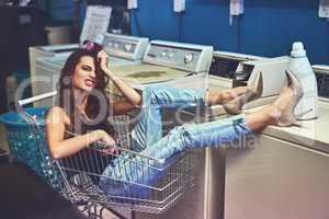 I have so much time on my hands today. Portrait of a attractive young woman seated inside of a shopping cart while resting her legs on a washing machine inside of a laundry room.