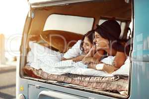 Anywhere is better together. Shot of two happy friends relaxing on a blanket in the true of their van on a road trip.