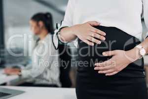 It looks like someone might arrive early. Cropped shot of an unrecognizable pregnant businesswoman standing and holding her stomach while a colleague works behind her.