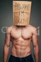 Beware what people think. Shot of an unrecognizable man wearing a paper bag saying beware Im ugly while standing against a grey background.