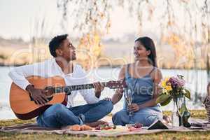 This song goes out to my special lady. Shot of a young man playing a guitar while on a picnic with his girlfriend at a lakeside.