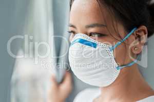 This crisis has induced wide reaching uncertainty. Shot of a young woman wearing a mask while looking out a window at home.