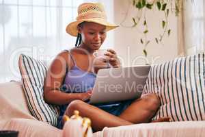 Theres no place like home. Shot of a young woman wearing a sunhat and using her laptop while having a coffee break on the sofa.