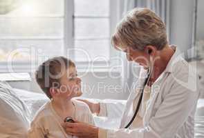 Take a deep breath for me. Shot of a doctor examine a little boy with a stethoscope in bed at home.