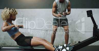 Beat the clock, become the best. Shot of a young woman exercising on a cable row machine with a fitness trainer in a gym.