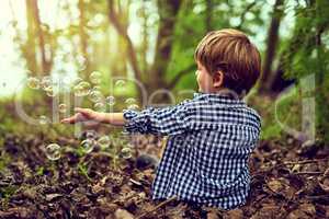 Fascinated by the magic of bubbles. Shot of a little boy playing with bubbles while sitting alone in the forest.