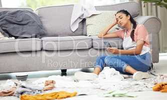 I need some help with these chores. Shot of a woman sitting on the floor with her cellphone in a messy living room.