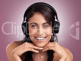 Where words fail music speaks. Shot of a beautiful young smiling woman with her hands rested on her chin while wearing headphones against a pink background.