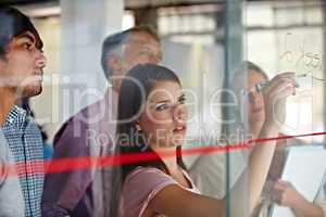 Explaining her ideas with a drawing. A young businesswoman writing down plans on a glass pane while her associates look on.