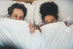 Guess what were hiding. High angle portrait of an affectionate young couple playfully covering themselves with a blanket in their bedroom at home.