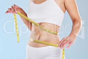 A woman using a measuring tape to measure her flat stomach against a isolated blue studio background. Closeup hands of a female taking waist measurements to track weight loss progress while on a diet