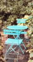 Two courtyard chairs and a blue metal table in a peaceful private home garden with green plants. Comfortable outdoor patio furniture set and seating in an empty, tranquil backyard with fresh flowers