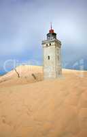 Lighthouse on a sand dune by the sea against a blue sky. Mysterious old tower alone in the desert. Deserted lighthouse isolated on beige sand. Peaceful and tranquil nature scene in Jutland, Denmark.