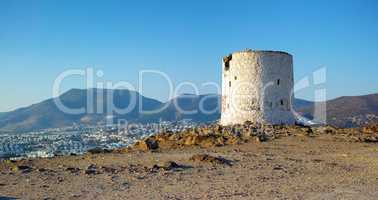Ruins of broken windmill on a hill overlooking a city surrounded by mountains. Crumbling walls of abandoned lighthouse tower. A weathered round white stone building. Architecture of an old structure