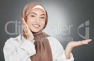 Portrait of glowing beautiful muslim woman isolated against grey studio background. Young woman wearing a hijab or headscarf showing flawless skin and holding her hand out for a product or treatment