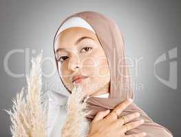 Studio portrait of one beautiful young muslim woman wearing brown headscarf posing with pampas wheat plant against grey background. Modest arab muslimah wearing makeup covered in traditional hijab