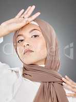 Modest female model wearing makeup with face covered in traditional hijab. Portrait of one beautiful young muslim woman wearing brown headscarf with hand on forehead against grey studio background.