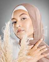 Studio portrait of one beautiful young muslim woman wearing brown headscarf posing with pampas wheat plant against grey background. Modest arab wearing natural makeup and covered in traditional hijab