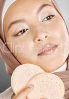 Beautiful muslim woman holding and using makeup sponges on her face against a grey studio background. Young female wearing an hijab and doing her beauty skincare routine. Taking care of healthy skin