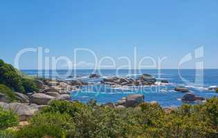 Landscape of rocks in the ocean on a hot summer day in Hout Bay, Cape Town. Calm waves splashing against rocky and shrubby coastline. Beautiful large shoreline rocks by the sea with lush vegetation