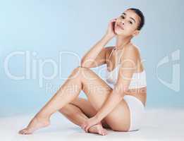 Portrait of a beautiful young woman posing in underwear while sitting in studio isolated against a blue background. Beauty shot of a confident and flawless model with soft, smooth and silky skin