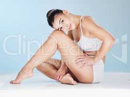 Portrait of a slim young latin woman sitting and posing in underwear against a blue background in a studio. Beautiful female with soft, smooth, and silky skin. Feeling sexy, fresh, and confident