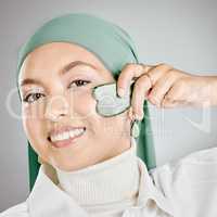 Portrait of a happy beautiful muslim woman using a gua sha on her face against a grey studio background. Young woman wearing a hijab and doing her skincare routine. Taking care of healthy skin