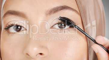 Closeup portrait of muslim woman applying mascara makeup to eyelashes. Headshot of stunning arab model doing her beauty routine against grey background. Face of beautiful woman extending her lashes